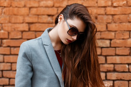 Beautiful young girl in sunglasses and gray coat looks down near brick wallの写真素材