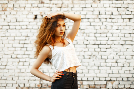 Beautiful young woman model in a white top shirt and vintage black jeans near a brick white wallの写真素材