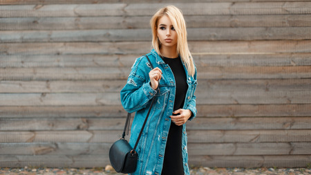 Beautiful young blonde woman with a handbag in a denim jacket near a wooden vintage wallの写真素材