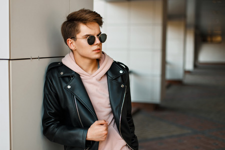 young handsome man in aviator sunglasses in a black leather jacket and a pink sweatshirt near a modern white wallの写真素材