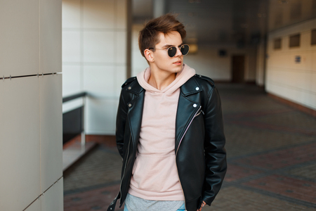 young handsome man in aviator sunglasses in a black leather jacket and a pink sweatshirt near a modern white wallの写真素材