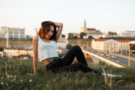 Beautiful young woman with a hairsyle sits on the grass on a city backgroundの写真素材