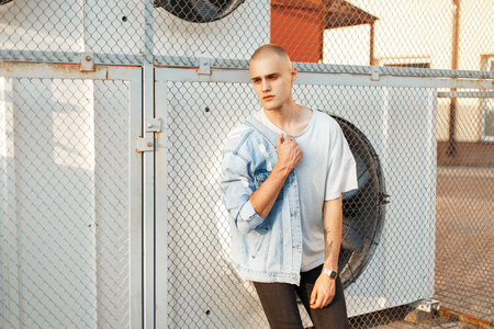 Handsome young bald man in a gray T-shirt and a denim jacket posing near the gridの写真素材