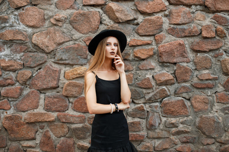 Beautiful young girl in black fashion clothes with a hat posing near a stone wallの写真素材