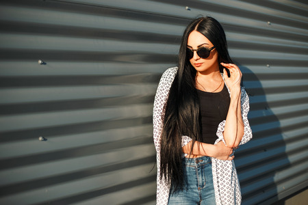 Stylish beautiful fashionable model of a girl with long hair wearing sunglasses in trendy clothes near a metal wall on a sunny dayの写真素材