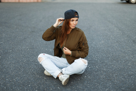 Beautiful stylish woman in a fashion military jacket with torn jeans sits on the asphaltの写真素材