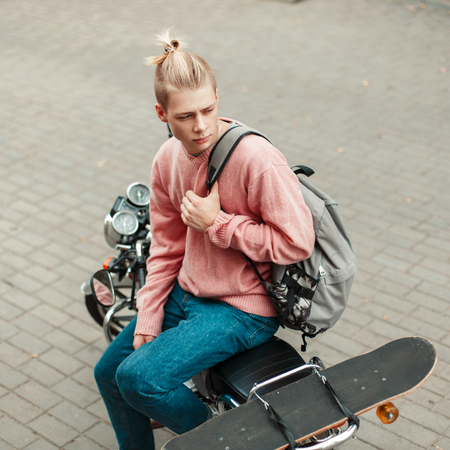 Handsome young man with a hairstyle in a pink sweater with a backpack and a skateboard sitting on a motorcycleの写真素材
