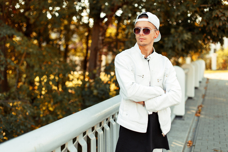 Handsome young man in sunglasses in a white baseball cap and autumn white jacket in the street on a sunny day near treesの写真素材