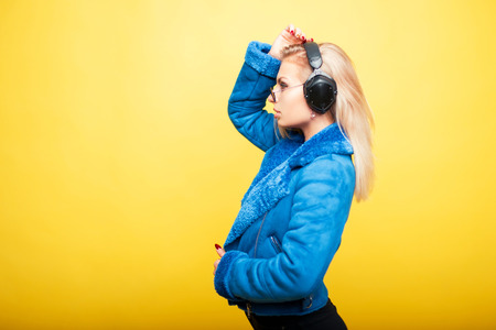 Beautiful young girl in fashionable sunglasses with headphones in a blue fashion coat on a yellow backgroundの写真素材