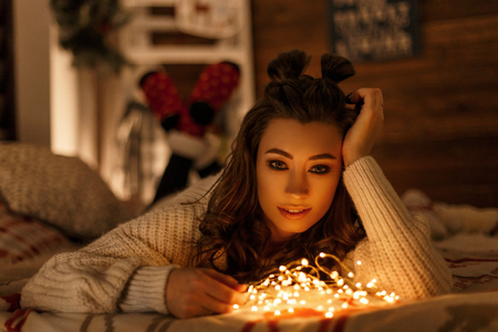 Beautiful young woman with festive lights in a vintage knitted sweater resting on a bed on Christmas Eveの写真素材