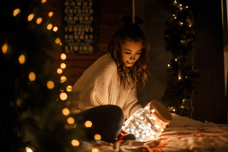 happy young girl in a vintage knitted sweater holds a magic jar with festive lights on the bedの写真素材