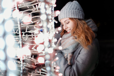 Happy beautiful girl in fashionable knitted winter clothes and a vintage hat with a scarf near the lights at nightの写真素材