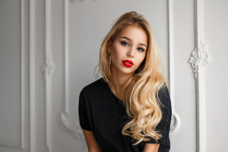 Beautiful young blonde model girl with red lips in a black t-shirt near a white elegant wallの写真素材