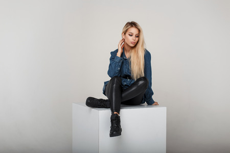 Pretty young blonde woman in fashion denim clothes in leather pants and black fashionable sneakers sits in studioの写真素材
