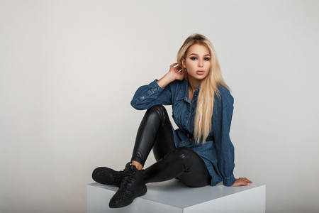 Beautiful young stylish woman in trendy denim clothes with black leather pants and black shoes sitting in studioの写真素材