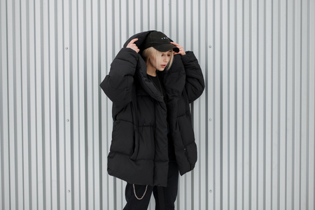 Beautiful young stylish woman in a trendy black cap and winter jacket near a metal wallの写真素材