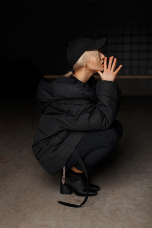 Stylish fashion young woman in a black fashionable hat with a black stylish winter jacket with boots sitting indoorsの写真素材