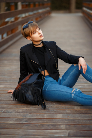 Fashionable young model in black stylish clothes with a jacket, jeans and boots with a leather bag sitting on a wooden bridge on the streetの写真素材