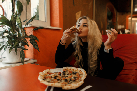 Stylish beautiful young woman eating pizza at a table in a cafeの写真素材