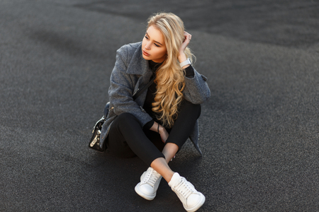 Beautiful fashionable young model woman in a stylish gray coat, black pants and white shoes sits on the asphaltの写真素材