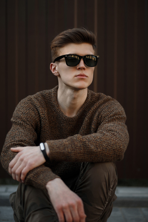 Handsome young American model man in sunglasses with a hairstyle in a fashionable pullover sits near a metal wallの写真素材