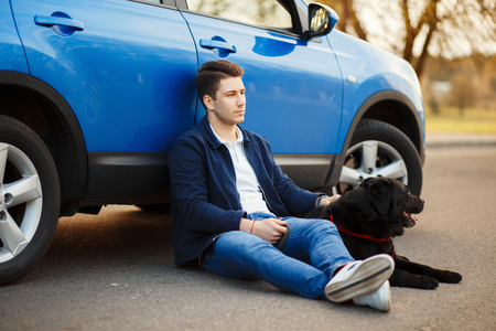 young handsome man in fashionable clothes sits near a car with a dog and resting on a spring dayの写真素材