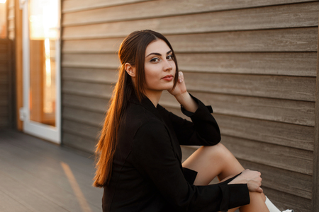 Pretty stylish model woman in a black elegant coat sits near a wooden wall at sunsetの写真素材