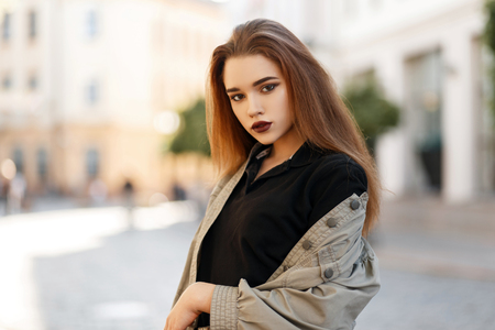Stylish pretty young woman with makeup in black polo shirt and fashion jacket posing outdoors on a sunny dayの写真素材