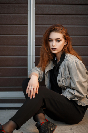 Beautiful young model woman in fashionable jacket and black T-shirt with pants sitting on the street near the gateの写真素材