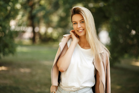 Happy young woman with a smile in a fashion pink jacket posing in the park on a summer dayの写真素材