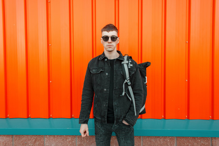 Handsome fashionable young model man with sunglasses in a black denim jacket with a backpack on the...