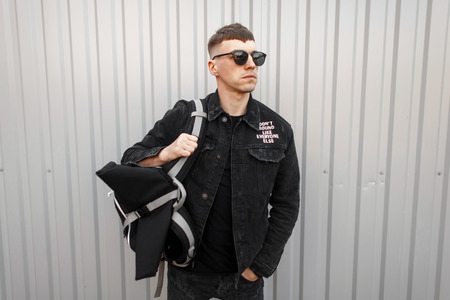 Handsome young man in a black denim jacket with a T-shirt in stylish sunglasses stands near a gray...