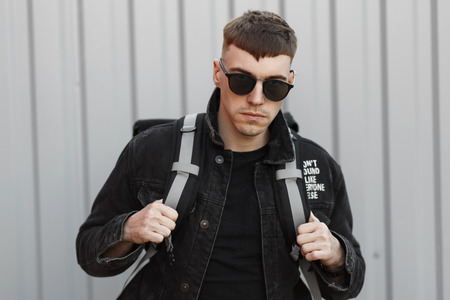 Stylish handsome man with a hairstyle with sunglasses in a fashionable denim black jacket with a bag...
