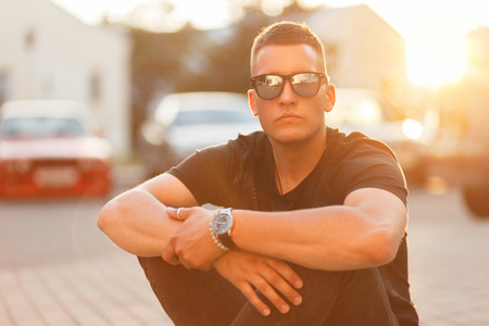 Stylish handsome young man with sunglasses in trendy black clothes sits asphalt at sunsetの写真素材