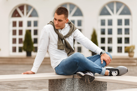 Fashionable handsome young man businessman in a white shirt with a sweater and jeans sitting on a bench in the cityの写真素材