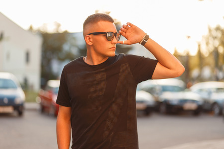 Fashionable handsome guy with a stylish hairstyle with glasses in a black T-shirt in the city at sunsetの写真素材