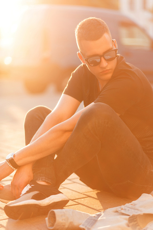 Fashionable handsome young man with a hairstyle in sunglasses and a black T-shirt with jeans with sneakers sits at sunsetの写真素材