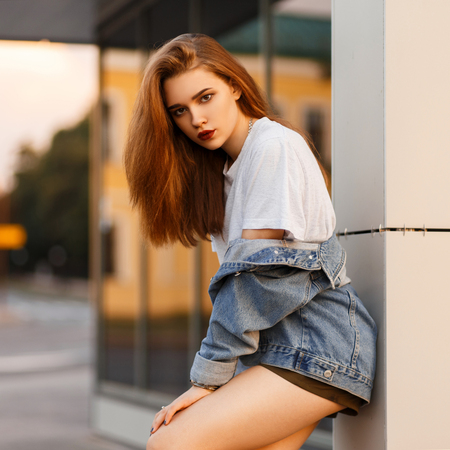 Stylish beautiful sexy model woman in trendy clothes with a jeans jacket posing outdoors near a buildingの写真素材