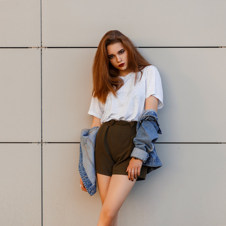 Beautiful fashionable young model woman in a denim jacket with a gray stylish t-shirt stands near a wall in the streetの写真素材