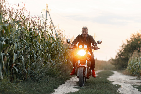 A brutal man in a military jacket rides a motorcycle with light in a cornfield