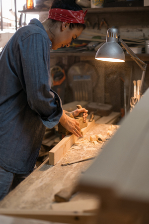Carpenter woman in denim clothing works in a wood workshop with a toolの写真素材