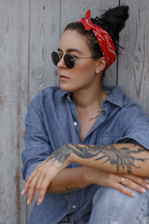 Pinup style. Stylish woman with a bandana and sunglasses in fashion jeans clothes sits near a wooden wall. Tattoo on the handの写真素材