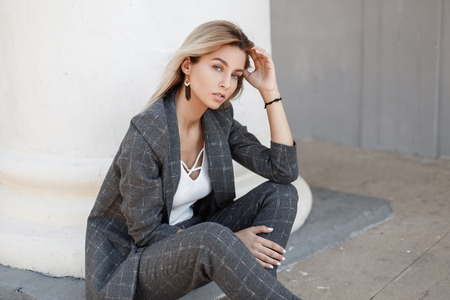 Young beautiful fashion model woman in stylish vintage gray suit sitting near white colognes on the...