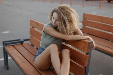 Stylish beautiful young woman model with curly hairstyle in casual clothes sitting on a wooden benchの写真素材