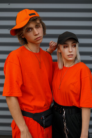 Fashionable young beautiful couple with caps in bright orange fashionable clothes near the metal gray wall. Fashion man and womanの写真素材