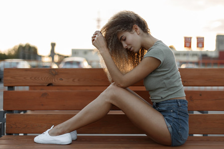 Sexy pretty young curly woman in denim shorts with white sneakers sitting on a benchの写真素材