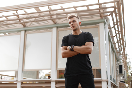 Young handsome fashionable american model man with hairstyle in black t-shirt on the street near a wooden buildingの写真素材