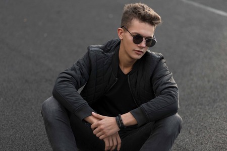 Fashionable hipster man in black stylish clothes sits on the streetの写真素材