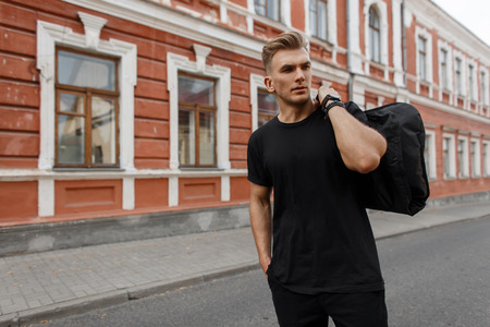 Handsome young stylish model man with hairstyle in black t-shirt with black bag on his shoulder walks on the streetの写真素材