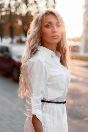 Fashionable pretty stylish young woman with natural makeup in a fashionable white shirt on the street at sunsetの写真素材